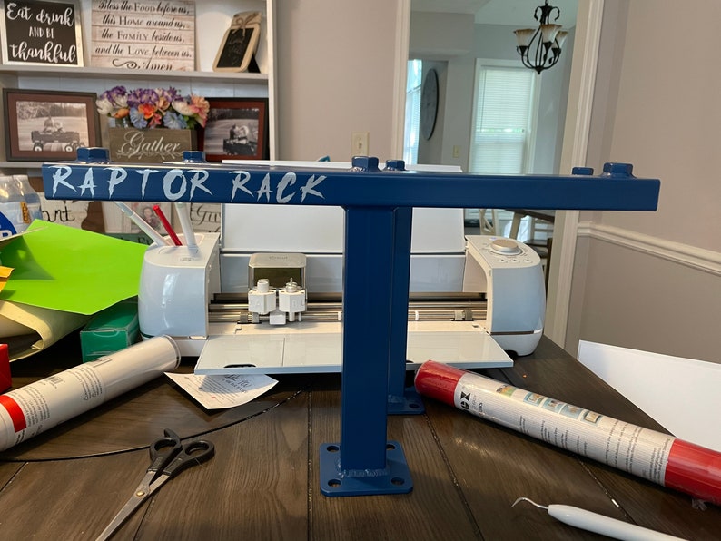 May include: A blue metal rack with the text "RAPTOR RACK" in white. The rack has a T-shaped design with a horizontal bar and a vertical support. The rack is on a wooden table, with a white cutting machine and craft supplies in the background.