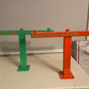 May include: Two metal racks, one green and one orange, are displayed. Each rack has a rectangular base with four holes, a vertical support, and a horizontal bar with the text "RAPTOR RACK" in a contrasting color. The racks are on a white surface.