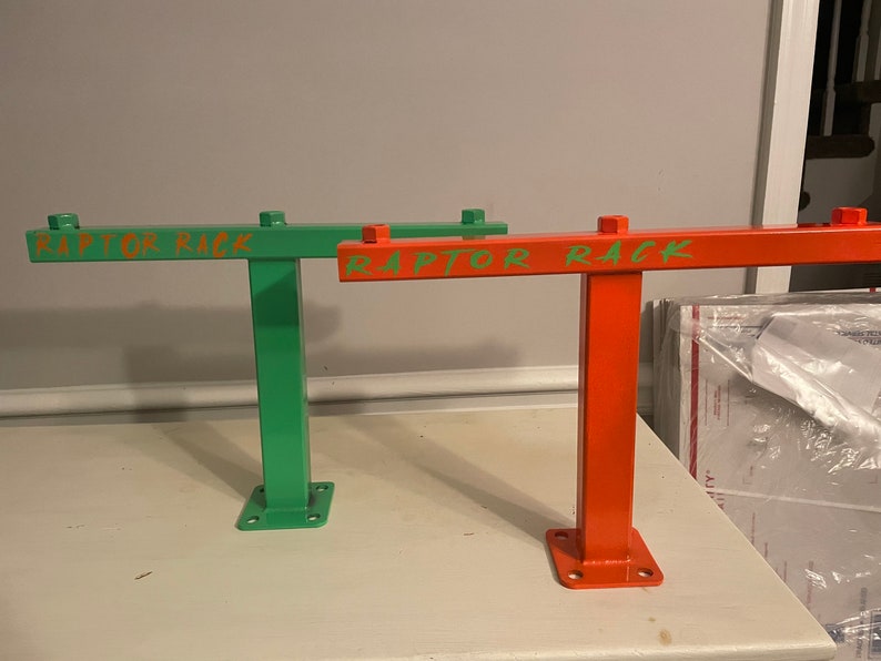 May include: Two metal racks, one green and one orange, are displayed. Each rack has a rectangular base with four holes, a vertical support, and a horizontal bar with the text "RAPTOR RACK" in a contrasting color. The racks are on a white surface.