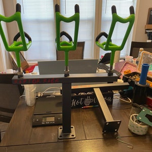 May include: A black metal fishing rod rack with three bright green rod holders. The text "RAPTOR RACK" is visible on the rack. The rack is on a dark wooden table, with other tools and items in the background.