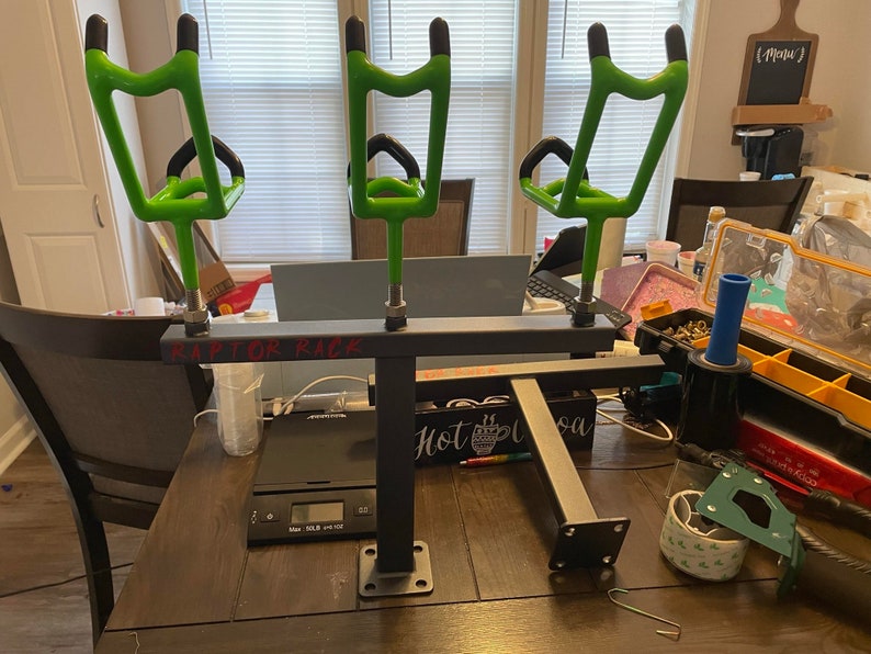 May include: A black metal fishing rod rack with three bright green rod holders. The text "RAPTOR RACK" is visible on the rack. The rack is on a dark wooden table, with other tools and items in the background.