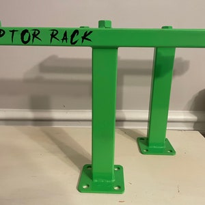 May include: Bright green metal rack with the text "RAPTOR RACK" in black. The rack has a rectangular frame with two vertical supports and a base with four holes for mounting. The rack is designed for heavy-duty use.