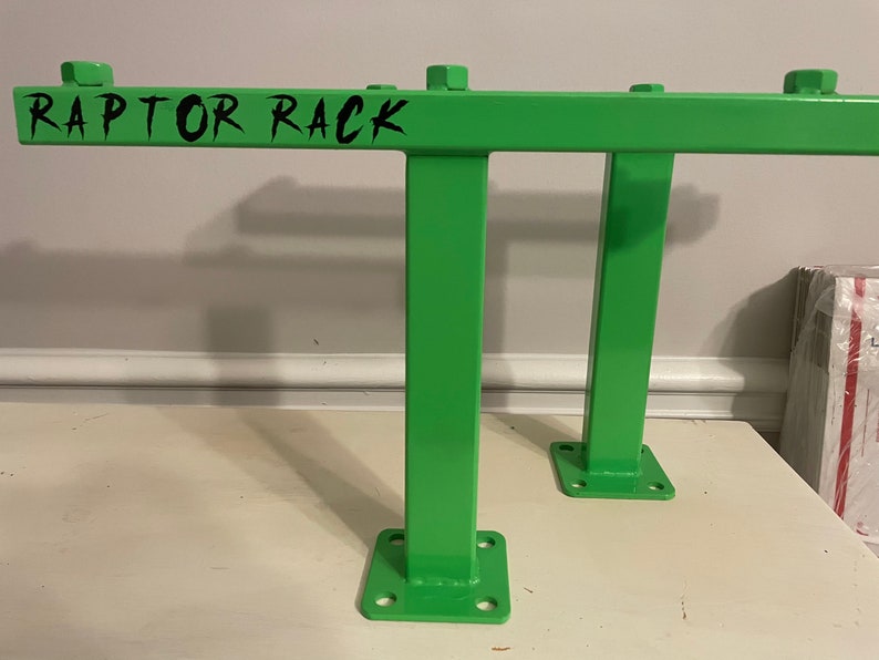 May include: Bright green metal rack with the text "RAPTOR RACK" in black. The rack has a rectangular frame with two vertical supports and a base with four holes for mounting. The rack is designed for heavy-duty use.