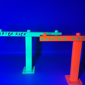 May include: Two metal Raptor Racks, one in bright green and the other in orange, are displayed against a blue background. Each rack has a horizontal bar with the text "RAPTOR RACK" in a contrasting color. The racks have a square base with bolt holes.