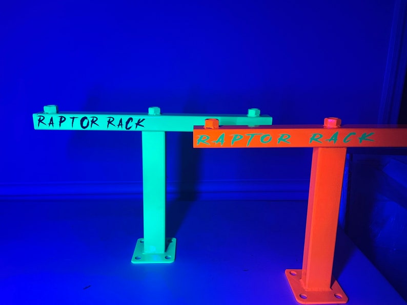 May include: Two metal Raptor Racks, one in bright green and the other in orange, are displayed against a blue background. Each rack has a horizontal bar with the text "RAPTOR RACK" in a contrasting color. The racks have a square base with bolt holes.