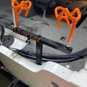 May include: A black metal Raptor Rack with two orange rod holders. The rack is mounted on a white boat. The text "RAPTOR RACK" is written in orange on the rack.