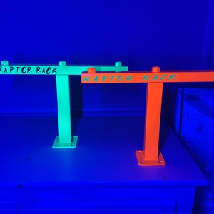 May include: Two metal Raptor Racks, one neon green and one bright orange, are displayed. Each rack has a horizontal bar with the words "RAPTOR RACK" in green. The racks are mounted on a square base and have bolts at the top.