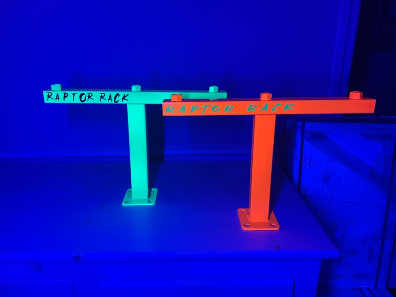 May include: Two metal Raptor Racks, one neon green and one bright orange, are displayed. Each rack has a horizontal bar with the words "RAPTOR RACK" in green. The racks are mounted on a square base and have bolts at the top.