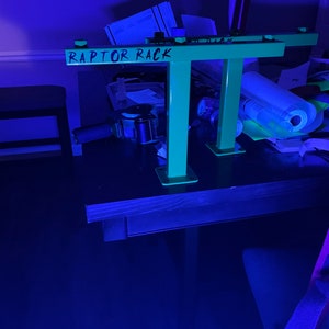 May include: A neon green metal rack with the text "RAPTOR RACK" in black. The rack has a rectangular top and two vertical supports. The supports are attached to square bases. The rack is on a dark surface.