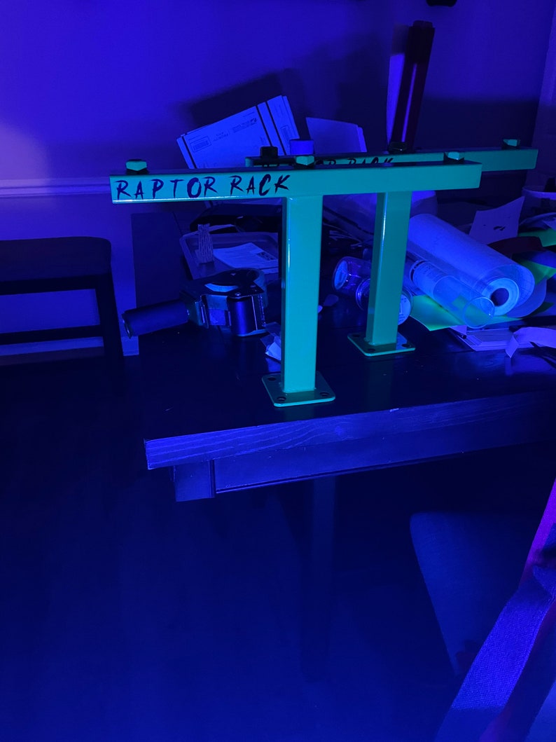 May include: A neon green metal rack with the text "RAPTOR RACK" in black. The rack has a rectangular top and two vertical supports. The supports are attached to square bases. The rack is on a dark surface.