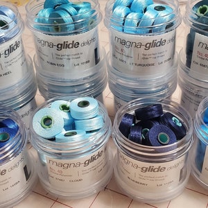 May include: A collection of small, clear plastic containers filled with spools of colorful thread. Each container is labeled "Magna-glide" and has a number and color name. The colors include: Tar Heel, Brown Egg, LT Turquoise, Persian, Cloud, Blue Berry, and Aqua.