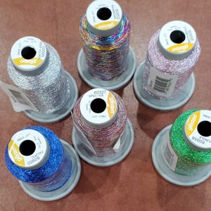 May include: Six spools of metallic thread in various colors, including silver, pink, green, blue, red, and rainbow. Each spool has a label with the brand name "glisten" and a product number.