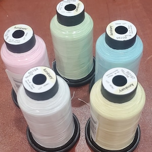May include: Five spools of thread in different colors: pink, white, green, blue, and yellow. Each spool has a black label with the brand name "luminary" and a product code. The thread is for sewing and crafting.