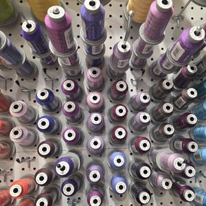 May include: A collection of colorful spools of thread, arranged on a pegboard. The spools are in shades of purple, pink, blue, and orange. The thread is wound around the spools, and the ends are visible. The pegboard is white.