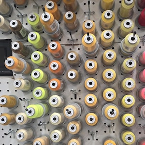 May include: A display of spools of thread in various shades of yellow, orange, and beige. The spools are mounted on a white pegboard, creating a visually organized and colorful arrangement. The thread is likely for sewing or crafting projects.