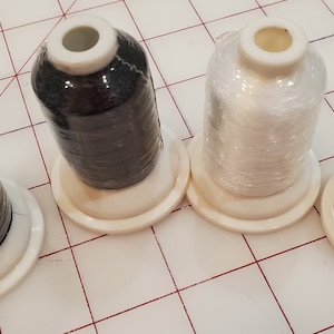 May include: Four spools of thread, two black and two white, on white plastic bobbins. The spools are wrapped in clear plastic.