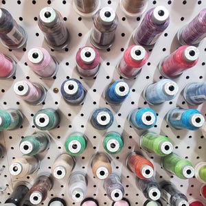 May include: A wall display of colorful spools of thread, organized in rows on a white pegboard. The spools are arranged in a variety of colors, including red, blue, green, and yellow.