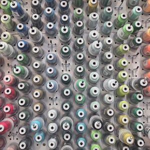May include: A close-up shot of a pegboard filled with numerous spools of thread. The spools are arranged in neat rows and columns, showcasing a wide array of colors, including greens, blues, yellows, and reds. The thread is wound around cylindrical spools.