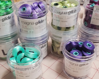 Peut inclure: Gros plan sur dix contenants en plastique transparent remplis de bobines de fil de couleur. Chaque contenant porte une étiquette indiquant "magna-glide" et comprend la couleur du fil, le numéro et d'autres détails. Les couleurs du fil comprennent le vert, le violet, le vert clair et le violet foncé.