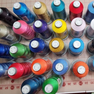 May include: A collection of 25 spools of embroidery thread in various colors, including blue, green, red, orange, yellow, purple, black, and white. The spools are arranged in a circular pattern on a gray surface with a ruler.