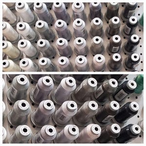 May include: A wall of spools of thread in various shades of gray, black, and white. The spools are arranged in rows on a white pegboard.