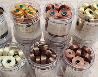 Peut inclure: Huit contenants en plastique transparent remplis de bobines de fil. Le fil est de différentes couleurs, notamment le brun, l'or et le beige. Les contenants sont étiquetés "Magna-glide" et portent le numéro "40".
