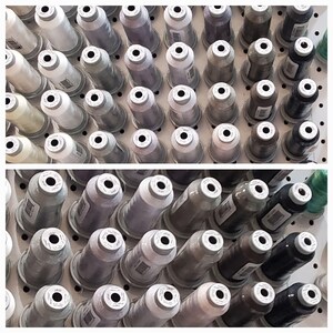 May include: Rows of thread spools in various shades of gray, white, and black are arranged on a white pegboard. The spools are cylindrical with a white top and a black hole in the center. The thread colors range from light to dark, creating a gradient effect.