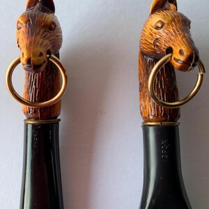 May include: Two brown wooden horse head bottle stoppers with gold rings. The stoppers are black on the bottom with the word "Japan" printed on them.