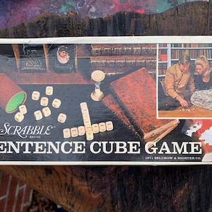 May include: A vintage Scrabble Sentence Cube Game box with a white background and a colorful image of a couple playing the game. The box features the Scrabble brand logo and the text "Sentence Cube Game" in bold black letters. The copyright date "1971 Selchow & Righter Co." is printed in smaller black letters below the game title.