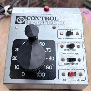May include: A silver Control Master II model train controller featuring a large black circular dial with numbers from 0 to 100. The unit includes black toggle switches for power, direction, momentum, and a red brake switch, all labeled on the metal face.
