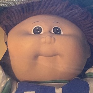 May include: A Cabbage Patch Kid doll with brown hair and a blue and green jersey. The doll has a light brown complexion and blue eyes.