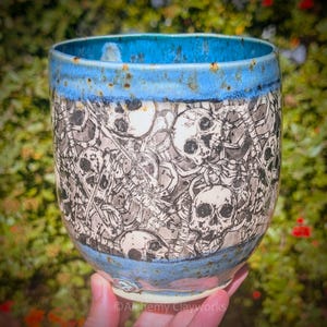 May include: A ceramic mug featuring a detailed black and white pattern of multiple skulls and skeletal bones across the body. The rim and the base of the mug are finished with a vibrant blue glaze that shows subtle brown speckling throughout.