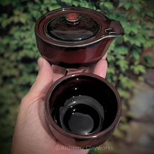 Pode incluir: Um pequeno bule de cerâmica vermelho-escuro com tampa a condizer e bico lateral, emparelhado com uma chávena de cerâmica vermelho-escura profunda. Ambas as peças apresentam um acabamento liso e brilhante com sombras pretas subtis e o texto Alchemy Clayworks.