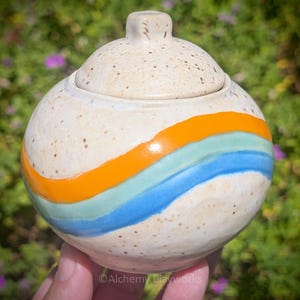 May include: A round ceramic lidded jar with a speckled cream-colored glaze. The exterior features three horizontal, wavy bands of color in bright orange, light mint green, and sky blue. The lid has a small, rounded knob handle on top.