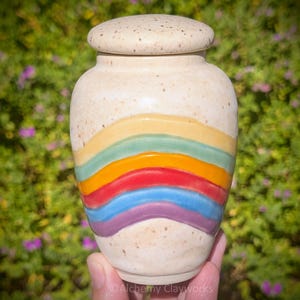 May include: A ceramic urn with a speckled cream-colored finish and a matching lid. The body features a series of horizontal, wavy bands in yellow, light green, orange, red, blue, and purple, creating a colorful rainbow-inspired pattern.