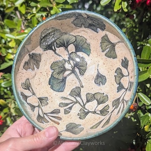 May include: A round ceramic bowl with a speckled beige interior featuring a dark green ginkgo leaf pattern. The bowl has a thick, dark teal rim and a smooth, glazed finish. The text Alchemy Clayworks is printed at the bottom of the bowl.