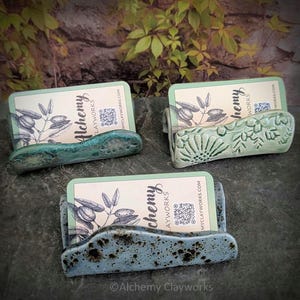 May include: Three ceramic business card holders with wavy edges. One is dark teal, one is light sage green with embossed floral patterns, and one is light blue with dark speckles. Each holds a card labelled Alchemy Clayworks with a QR code.