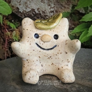 May include: A whimsical, handmade ceramic figure with a smiling face, arms outstretched, and small feet. The figure is off-white with brown speckles, a small tan nose, and a green leaf on its head. The eyes are dark blue. The Wildlings Clayworks logo is at the bottom.