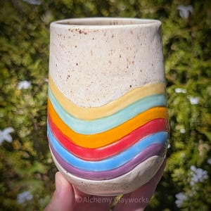 Op de afbeelding: Een keramische beker met een gespikkeld crèmekleurig bovendeel en een reeks van vijf horizontale, golvende, verhoogde banden in geel, lichtblauw, oranje, rood en paars over het onderste gedeelte. Het oppervlak heeft een getextureerde, matte afwerking.