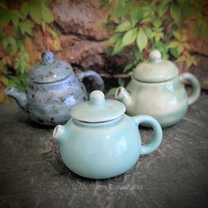 May include: Three small ceramic teapots in shades of blue and green. The front teapot is a light turquoise color, with a rounded body, a small handle, and a lid. The other two teapots are in the background, one blue and one green. The teapots are on a dark surface.