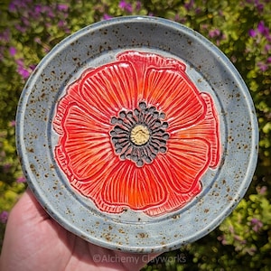 May include: A round ceramic plate featuring a large, embossed red poppy flower at its center. The plate has a speckled light blue-gray glaze, and the poppy has a textured yellow center with dark stamens. The text Alchemy Clayworks is visible.