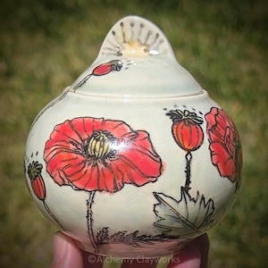 May include: A round ceramic lidded jar with a cream-colored glaze, featuring hand-painted red poppy flowers, green leaves, and black outlines. The lid has a pointed handle with a sunburst design. The text Alchemy Clayworks is visible.