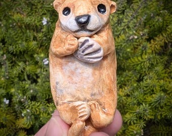 May include: A small, hand-sculpted clay otter figurine with a light brown textured finish, dark eyes, and a black nose. The otter is depicted in a lying position, holding a small, grey, textured shell-like object against its chest with its paws.