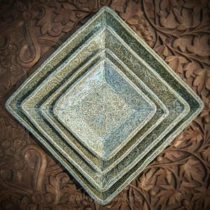 May include: A set of four square, nested serving dishes with a textured, floral pattern. The dishes are in shades of green and blue, arranged in a diamond shape. The background is a carved wooden surface.