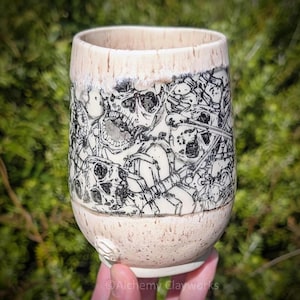 May include: A ceramic tumbler with a cream-colored speckled glaze featuring a detailed black ink illustration of multiple skulls and skeletal remains wrapped around the exterior. The rim has a smooth, lighter drip-style glaze finish.