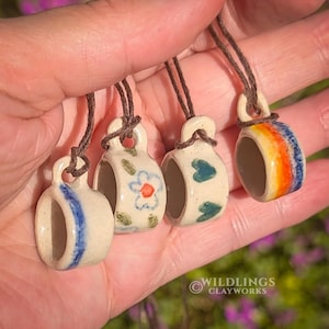 May include: Four small ceramic cup pendants with brown cords. Each cup is hand-painted with unique designs: blue stripes, a floral pattern, green hearts, and a rainbow. The pendants are held in a hand, showcasing the details and craftsmanship.