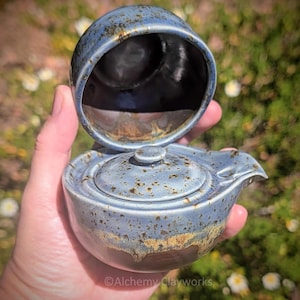 May include: A ceramic teapot with a lid, featuring a blue glaze with dark brown speckles and a smooth, glossy finish. The teapot has a small spout and a rounded body, with the text Alchemy Clayworks printed at the bottom of the image.