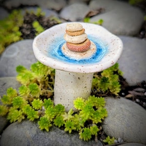 May include: A ceramic mushroom-shaped bird bath with a speckled white base and a shallow dish. The dish features a blue water design and holds a stack of small, round stones. Green plants and gray stones surround the bird bath.