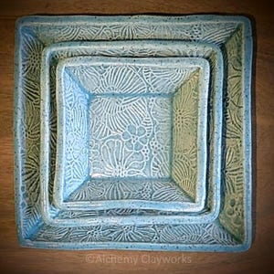 May include: A set of three nested square ceramic bowls in a light blue speckled glaze. Each bowl features a debossed floral pattern across the interior and rim. The text Alchemy Clayworks is printed in white at the bottom of the image.