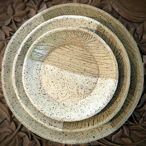 May include: A stack of three handmade ceramic plates in varying sizes. The plates feature speckled white and tan surfaces with textured wood grain and speckled designs. The plates are arranged on a carved wooden surface.